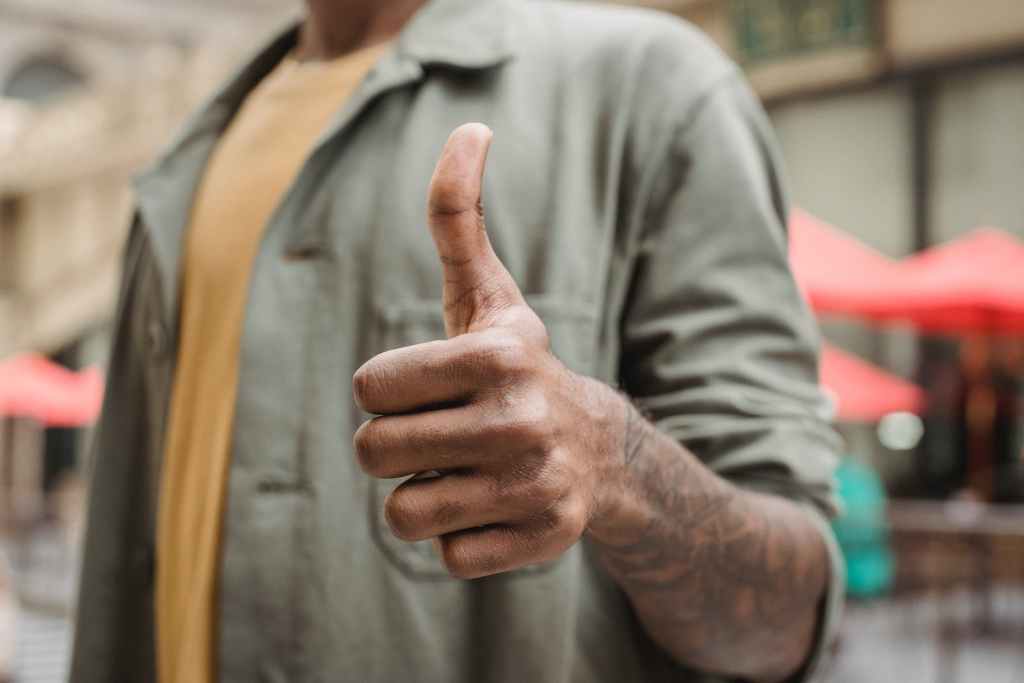 Crop faceless young black male in casual outfit standing on city street and showing thumb up gesture