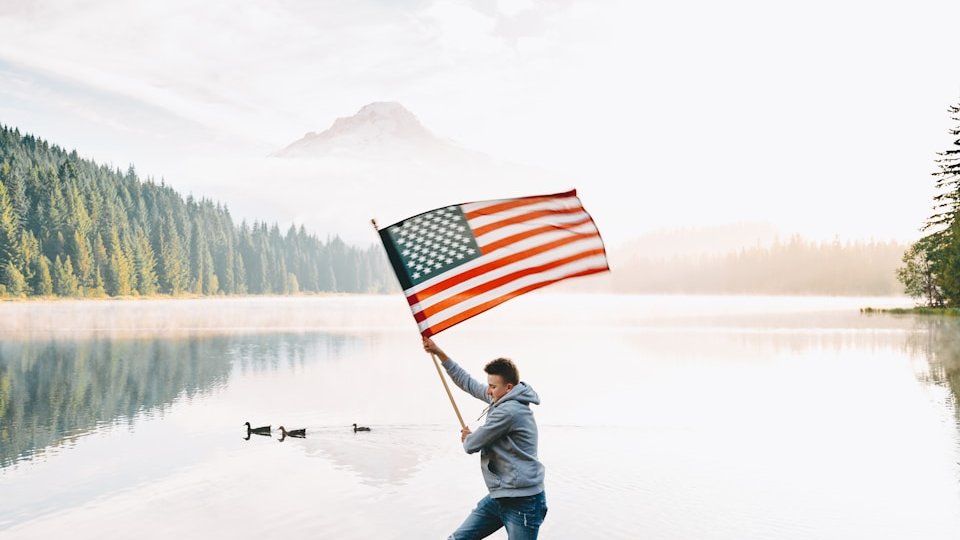 person holding us a flag standing on rock near lake during daytime