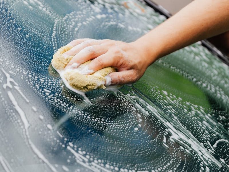 Close-up of a hand washing a car windshield with a soapy sponge, detailing care.